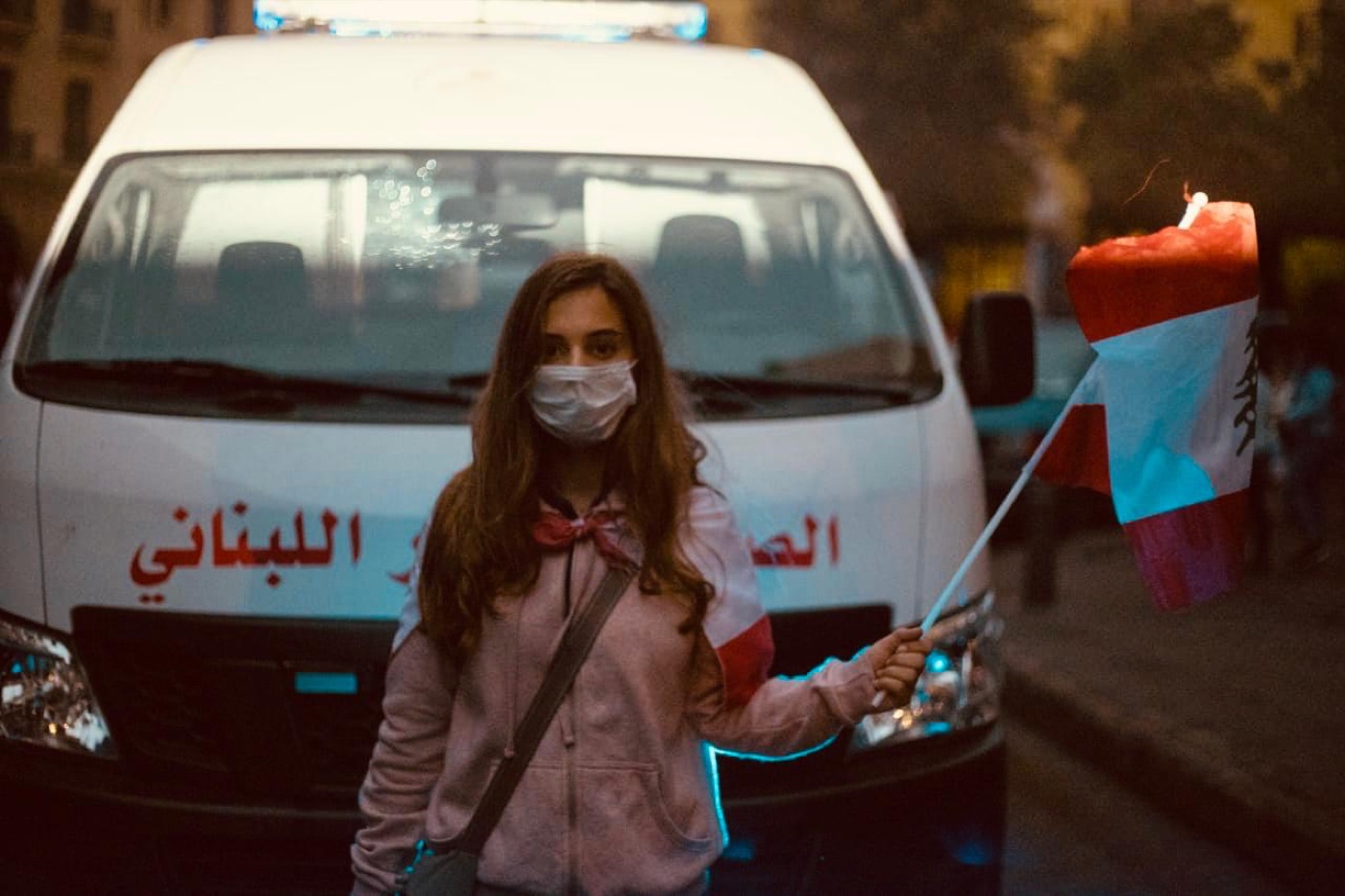 Myriam in a face mask holding a Lebanese flag in front of a Red Cross ambulance